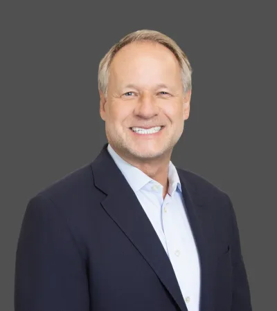 Smiling middle-aged man with light hair in a navy blazer and white shirt against a gray background