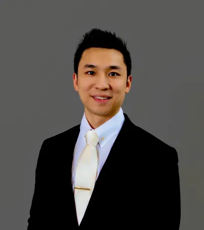 Professional young man in black suit, white shirt, and white tie smiling against a gray background.