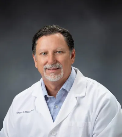 Middle-aged male doctor in a white coat with a light blue shirt, smiling against a gray background.