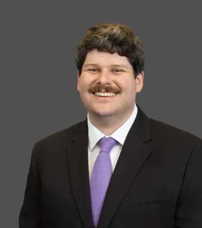 Smiling man with dark hair and mustache wearing black suit and purple tie on gray background