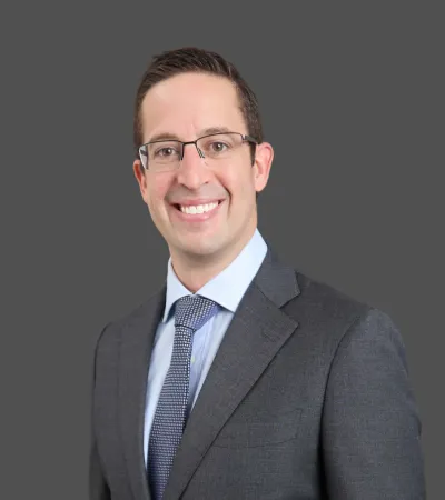 Professional man in glasses wearing a dark suit, light blue shirt, and patterned tie smiling against gray background