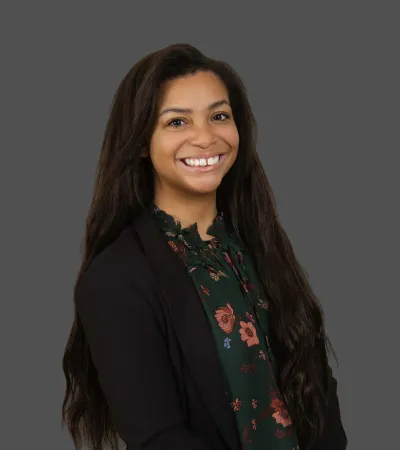 Smiling woman with long hair wearing floral blouse and black blazer against gray background
