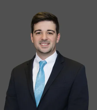 Smiling young man in a dark suit and blue tie against a plain gray background.