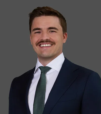 Smiling man with mustache wearing a dark suit, white shirt, and green tie against a gray background