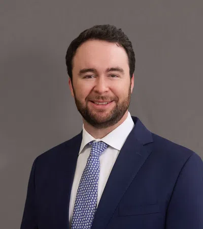 Smiling man in a navy blue suit and patterned tie against a gray studio background