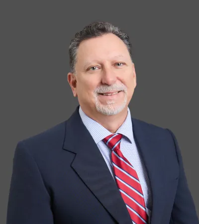 Middle-aged man in navy suit and red striped tie smiling against a gray background portrait.