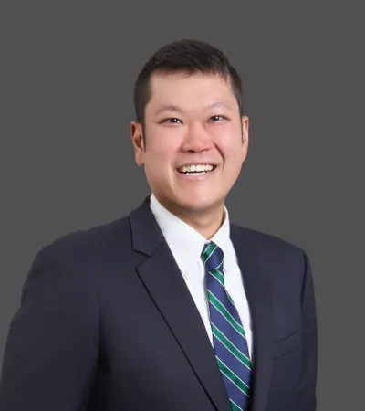 Smiling man in a dark suit and striped tie against a plain gray background.