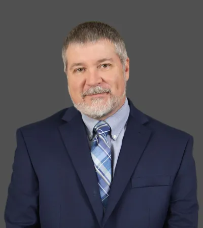 Middle-aged man with gray hair and beard in navy suit and blue plaid tie against gray background