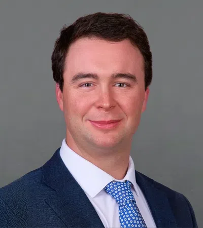 Professional headshot of a man in a dark blue suit and patterned blue tie against a gray background