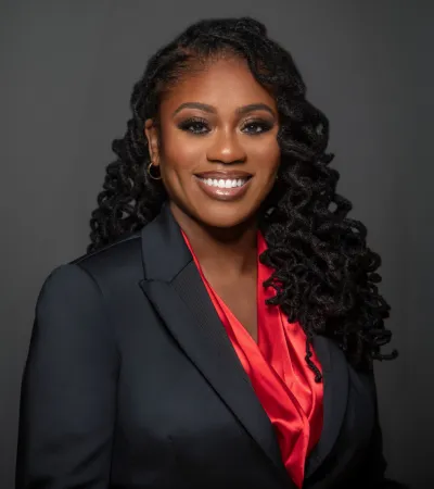 Smiling African American woman with curly hair wearing a black blazer and red blouse against a gray background