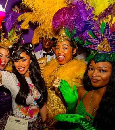 Group of people in colorful Mardi Gras costumes and feathers celebrating with drinks at a lively party.