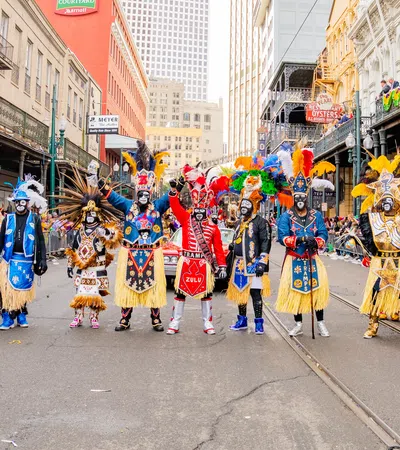 A colorful street parade with participants in feathered costumes and painted faces in an urban setting.