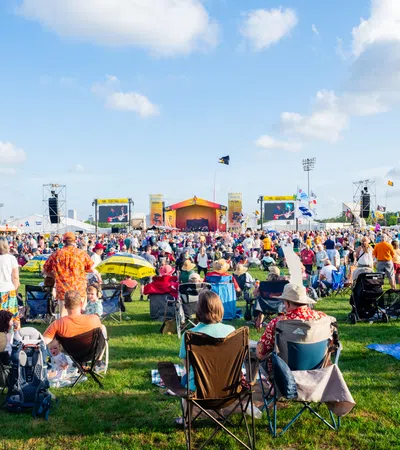 Large outdoor music festival crowd seated and standing on grass watching performances under blue sky with clouds.