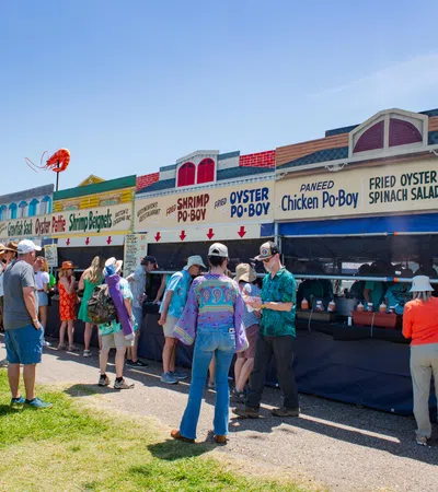 People lined up at outdoor food stalls serving seafood dishes like shrimp po'boy and fried oyster on a sunny day.