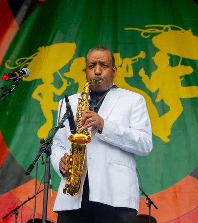 Saxophonist in white jacket performing on stage with colorful backdrop featuring silhouettes of musicians.