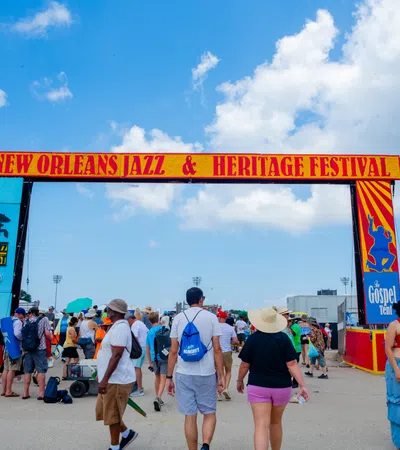 Crowd walking under New Orleans Jazz & Heritage Festival entrance arch on a sunny day with blue sky.