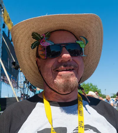 Smiling man wearing a straw hat and novelty sunglasses at an outdoor festival on a sunny day