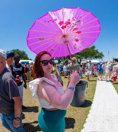 Woman holding a pink umbrella at a sunny outdoor festival with crowds and tents in the background