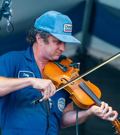 Musician wearing blue cap and shirt playing a violin on stage under a microphone.