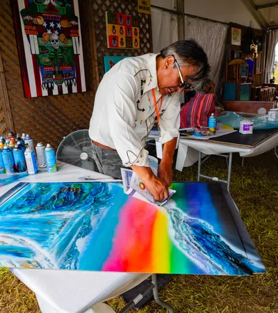 Artist creating a vibrant rainbow-themed painting indoors surrounded by spray paint cans and artwork.