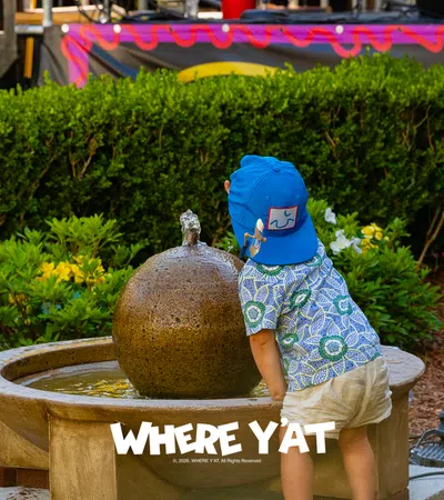 Toddler in blue hat and patterned shirt playing with water fountain in a park setting.