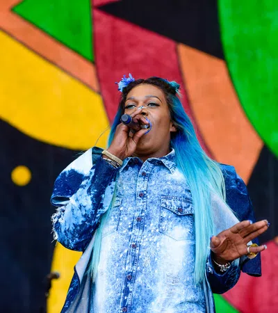 Female singer with blue ombre hair performing on stage with colorful abstract background art.