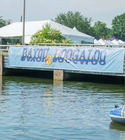 People in an inflatable boat on a bayou under a bridge with a Bayou Boogaloo festival banner.