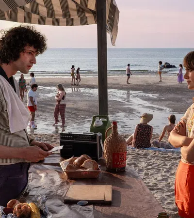 Young man and woman talking at a beachside stand with bread, drinks, and people walking along the shore at sunset.