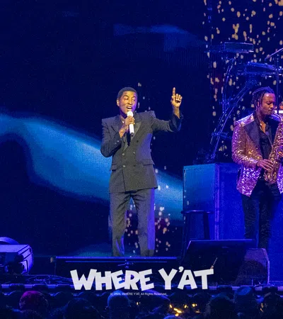 Singer performing on stage with saxophonist and DJ, vibrant lighting and audience visible in foreground