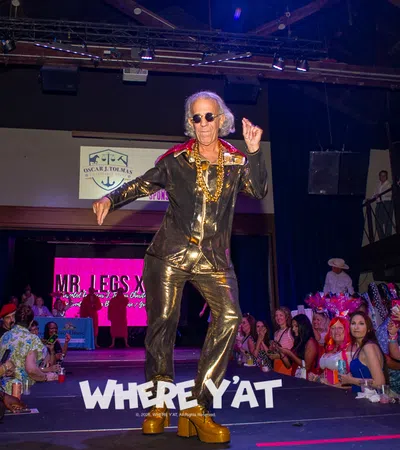 Older man in shiny gold outfit and platform shoes dancing on runway with cheering audience at event