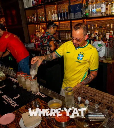 Bartender in Brazil soccer jersey preparing multiple drinks at a busy bar with diverse bottles behind the counter