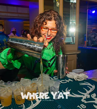 Bartender with curly hair and orange glasses pouring drinks into plastic cups at a lively bar.