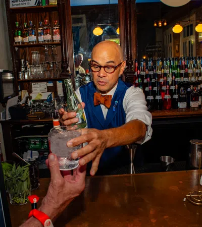 Bartender in a bow tie serving a cocktail at a wooden bar counter with bottles and glassware behind him