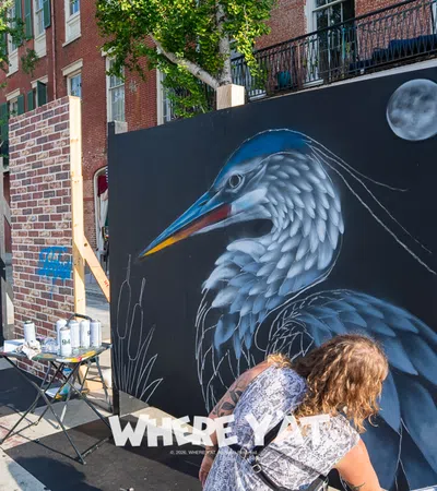 Street artist painting a large heron mural on black panels during an outdoor urban art event in the daytime.