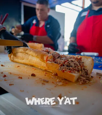Long sandwich being sliced, stuffed with shredded meat, chefs in red aprons working in background.