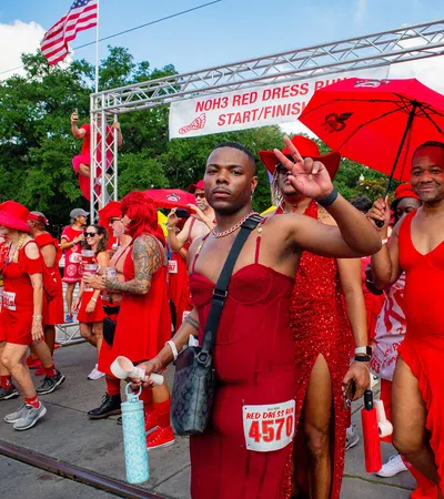 Participants dressed in red outfits posing at the NOH3 Red Dress Run start/finish line under a bright sky.