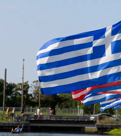 Greek flags waving alongside American flags by a riverside with people and greenery under a clear sky.