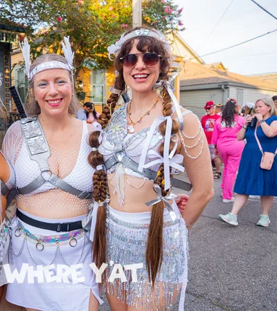 Three women in futuristic silver festival costumes posing outdoors at a sunny street event with a festive crowd.