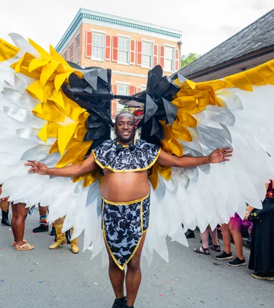 Man wearing large white, gold, and black wings costume at colorful outdoor street festival parade.