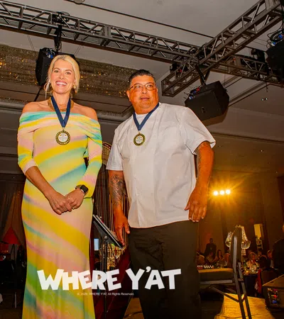 Two award recipients wearing medals stand on stage at a formal event with audience in background.