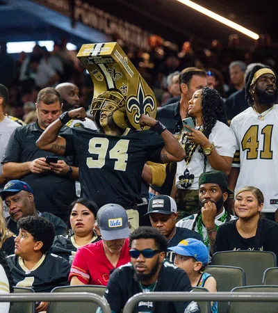 Excited fans at football game, some wearing New Orleans Saints jerseys and merchandise in stadium seats.
