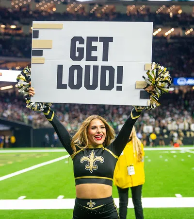 Cheerleader in New Orleans Saints uniform holding a GET LOUD! sign on football field with crowd behind
