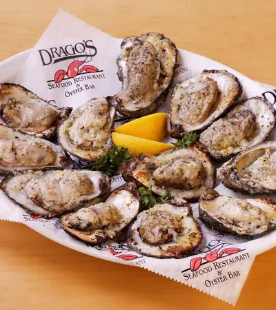 Grilled oysters on a white plate with lemon wedges, served on branded paper from Drago's Seafood Restaurant.