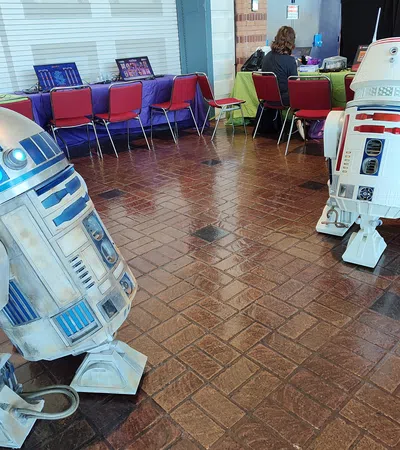 Two detailed Star Wars droids R2-D2 and an R2 variant stand on a tiled floor in a convention setting with laptops and chairs.