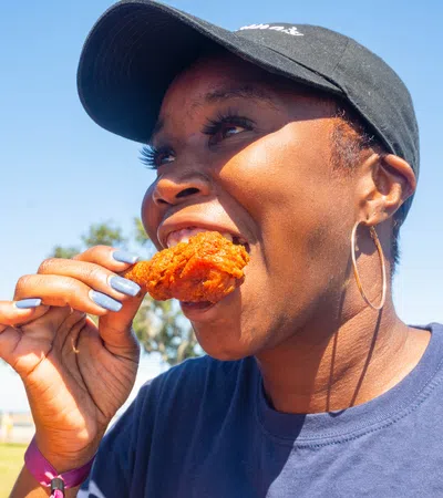 Smiling woman wearing a black cap and hoop earrings eating fried chicken outdoors on a sunny day