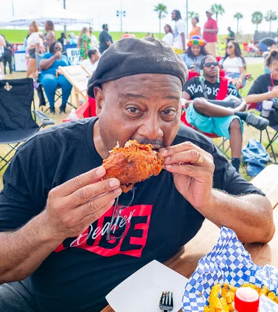 Man in black hat eating fried chicken at outdoor event with people and tents in the background