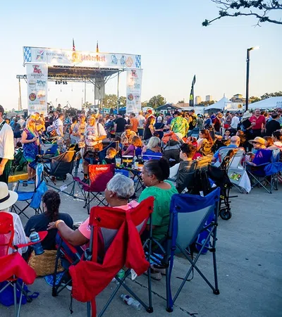 Crowd enjoying an outdoor music festival with people sitting on chairs and socializing near the stage at sunset.