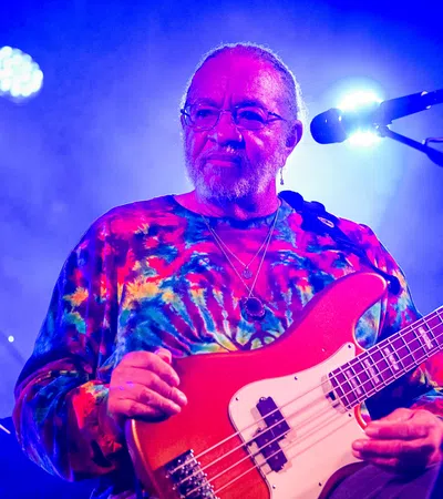 Musician wearing a colorful tie-dye shirt playing red electric bass guitar on stage with blue lighting.