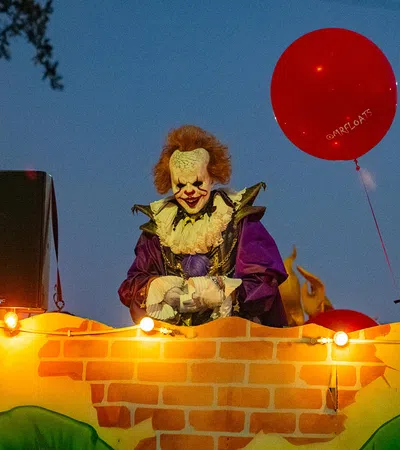 Costumed clown with red hair and white face paint holding red balloons at night during parade float event.