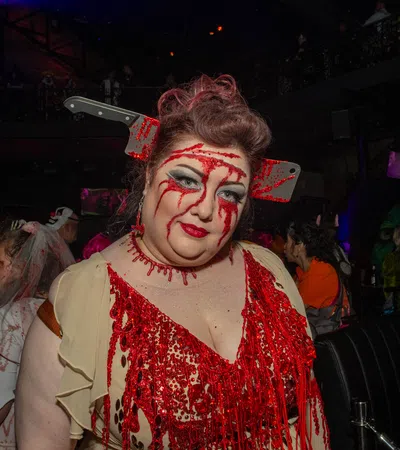 Woman in bloody costume with fake cleaver through head at a Halloween party with crowd in background